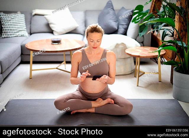 Pregnancy and technoloy supported healthy lifestyle concept. Cheerful happy pregnant woman using smart phone application while exercising on yoga mat on living...