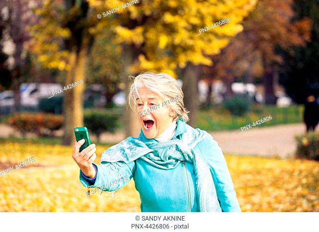 View of a smilling and espressive pretty senior woman looking at his phone on yellow leaves in front of a trees with autumnal colors