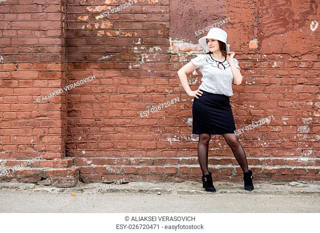 Woman in a white hat, blouse and black skirt, posing against the backdrop of an old vintage brown brick wall. Female portrait in a retro style