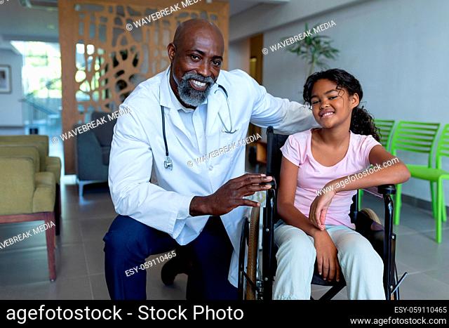 Portrait of smiling african american male doctor and mixed race girl in wheelchair in hospital foyer