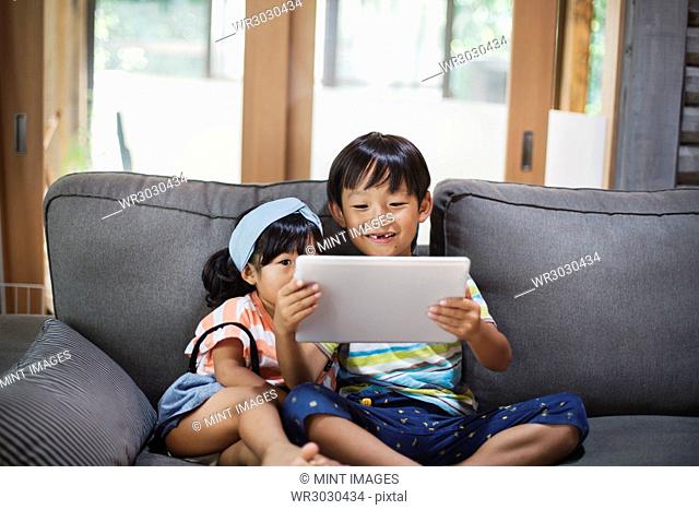 Boy and young girl with black hair sitting on a grey sofa, looking at digital tablet