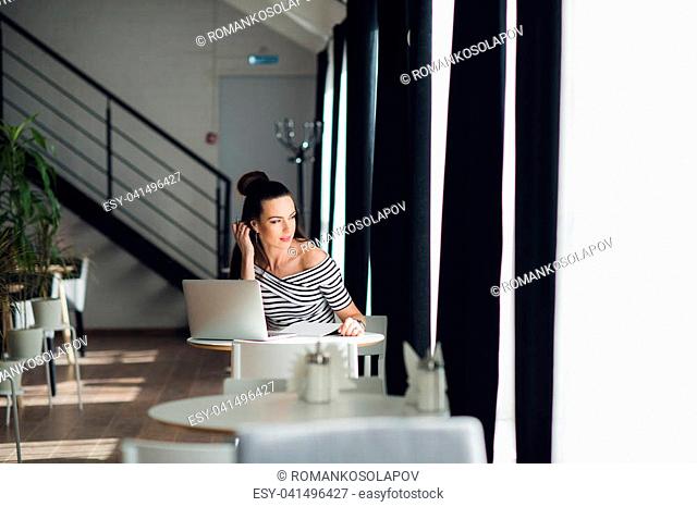 Looking for inspiration. Young beautiful pensive woman making some notes and looking through window while sitting in chair at her working place