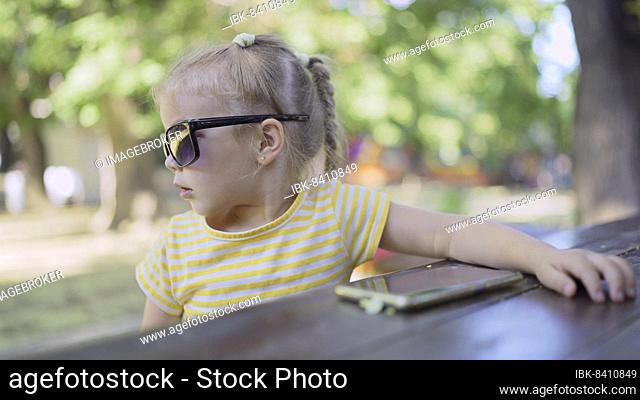 Cute little girl in moms sunglasses listens to music on a mobile phone and sings along. Close-up portrait of child girl sitting in sunglasses on city park and...