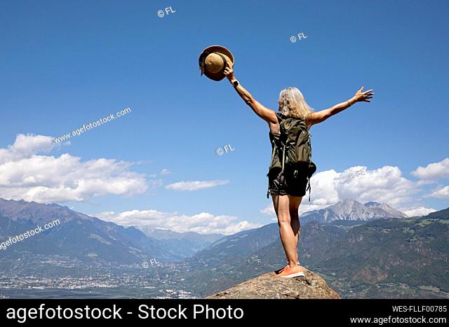 Carefree senior woman with arms raised standing on rock in front of mountains