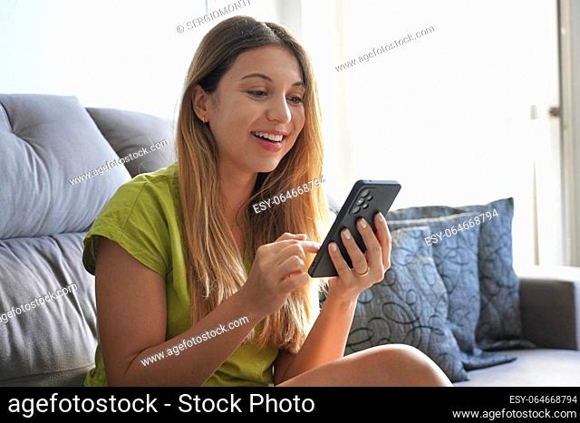 Happy lively girl resting on cozy sofa with smartphone, share messages to friend in social networks smile enjoy free time on internet