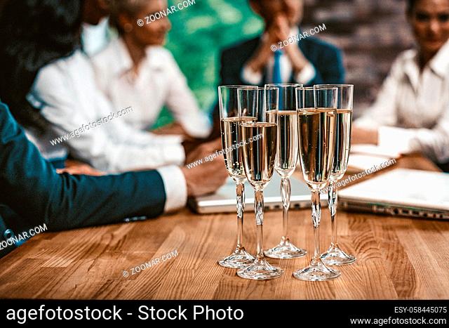 Five Glasses With Alkohol Are Lokated On Wooden Table In Office. Selective Focus On Wineglasses With Champagne, Meeting Of Business People on Blured Background