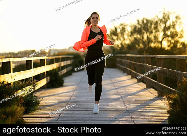 Smiling young woman running on footbridge against clear sky during sunset