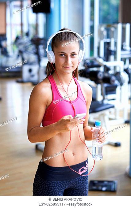 young healthy woman drinking water in fitness gym while sitting on pilates ball and listening music on headphones from smartphone