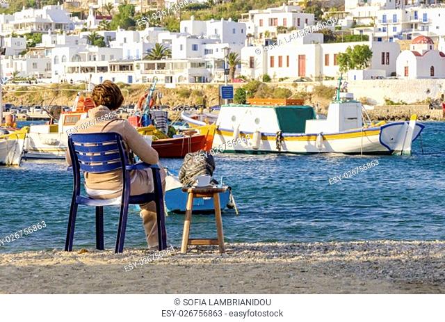 The coastline of Chora in Mykonos, Greece. A woman stting on the golden sand beach having coffee and gazing the view, typical greek island boats and whitewashed...
