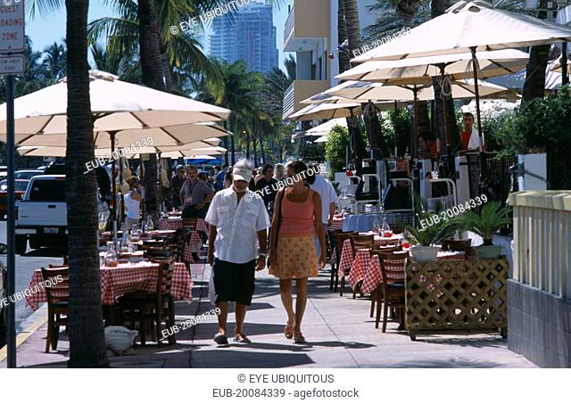 South Beach. Ocean Drive. A man and woman walking past a sidewalk cafe restaurant with table and chairs under sun umbrellas