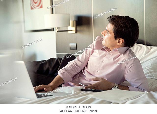 Businessman lying on hotel bed with cell phone and laptop computer, looking away in thought