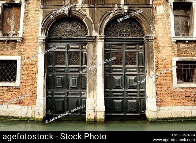 Wooden doors painted green in a red brick facade with wrought iron grilles at the sides in Venice, with canal water up to the door