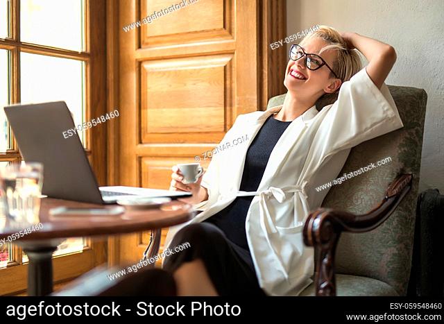 Dreamy blonde girl in glasses sits on the armchair at the table with a laptop in the cafe. She leans on armchair with a smile and holds a cup in right hand