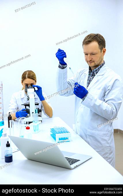 Male scientist analyzes liquid in beaker, female researcherworking with microscope, colleagues investigating medical sample together, conducting experiments