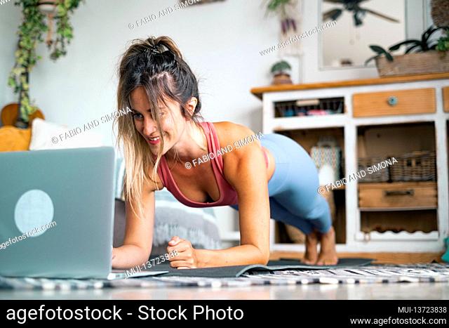 Young woman exercising at home doing push ups and looking at her personal laptop computer to learn or teach workout - content creator business free healthy...