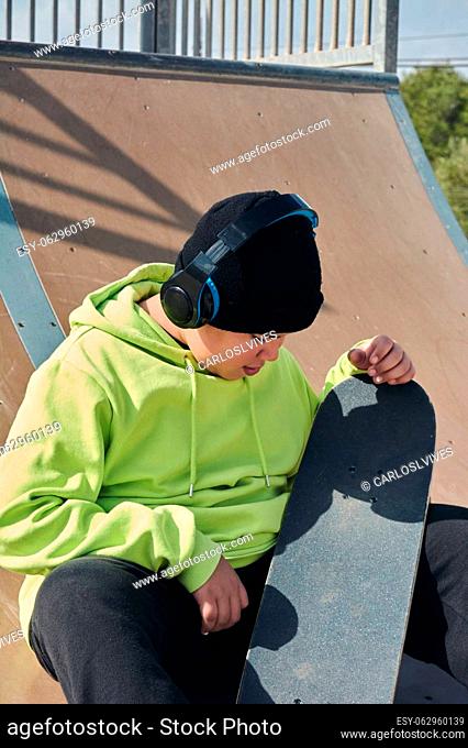 young, teenager, with a skateboard, thinking, on a rink, skateboarding, wearing headphones, green sweatshirt, black hat, swinging on a sunny day