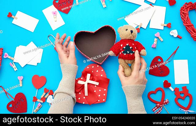 female hands holding a small brown teddy bear, on a blue background an open empty heart-shaped box, top view, holiday Valentine's Day