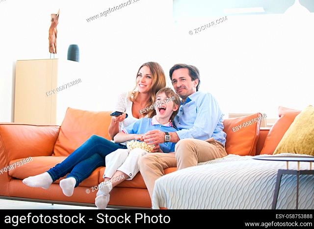 Beautiful young parents and their son are watching TV, eating popcorn and smiling while sitting on couch at home