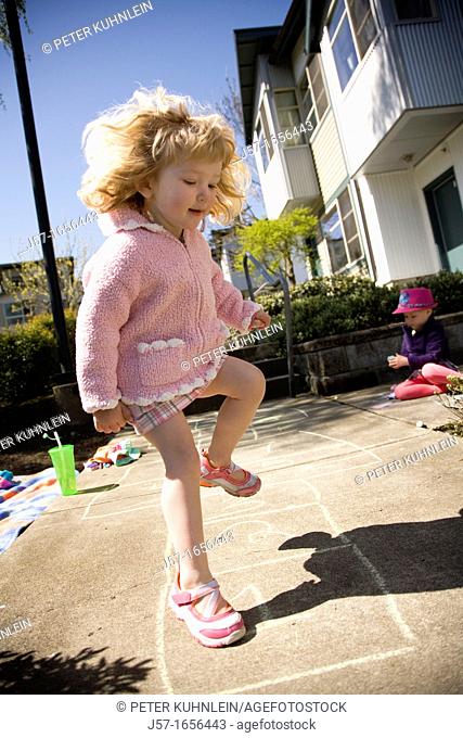 Young girl playing hopscotch infront of her appartment in the sun. Sister playing on the sidewalk behind her