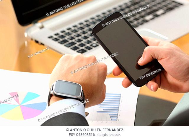 Businessman hands synchronizing on line a smart watch and phone sitting in a desktop at office with a computer in the background