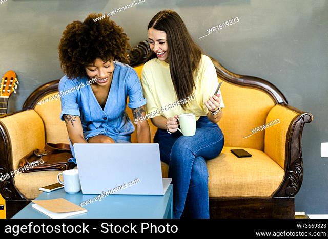 Cheerful women using laptop while sitting on sofa in modern cafe