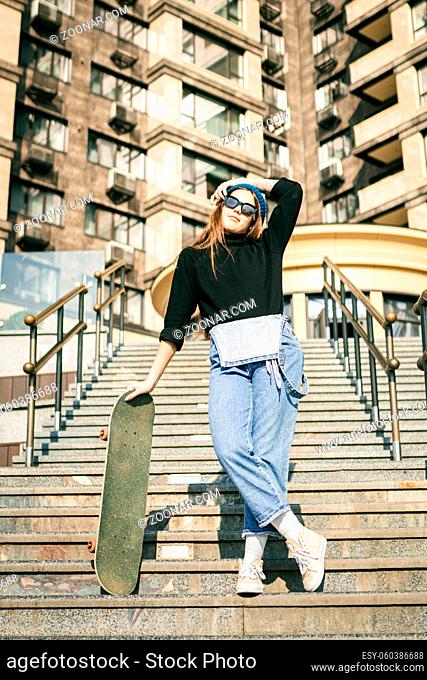 Stylishly dressed woman in blue denim jumpsuit posing with skateboard. Street photo. Portrait of girl holding skateboard. Lifestyle, youth concept