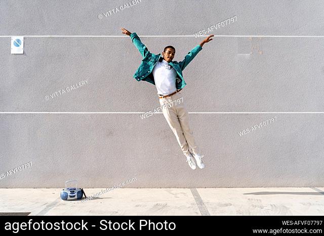 Young man with radio on footpath jumping against gray wall