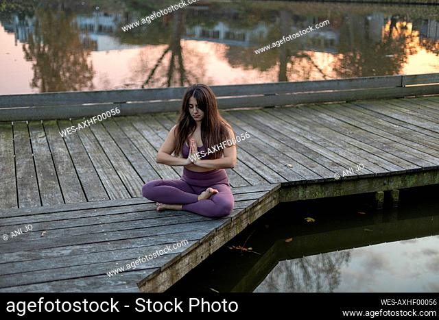Woman with long hair meditating while sitting on pier over lake at sunset