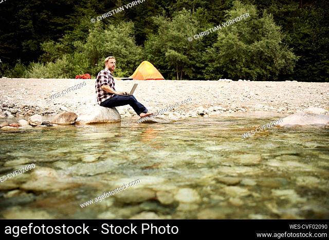 Businessman with laptop sitting on rock at riverbank