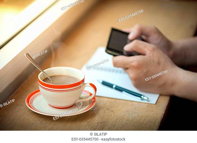 Male hand using smart phone on wood bar beside window with notebook and coffee cup in cafe in morning time