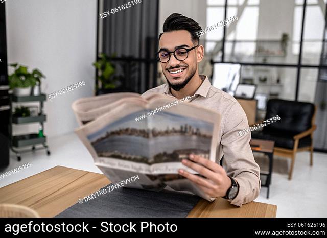 Morning newspaper. Joyful successful young bearded man in glasses reading newspaper sitting at table in cozy room