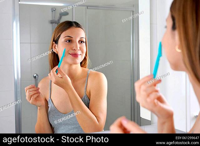 Facial hair removal. Beautiful young woman shaving her face by razor at home. Pretty woman using shaver on bathroom