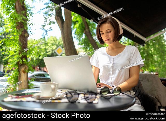 Young female professional using laptop while listening music through headphones sitting at cafe
