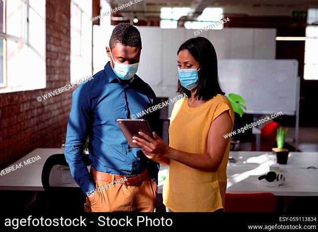 Diverse male and female colleague wearing face masks standing in office looking at tablet together
