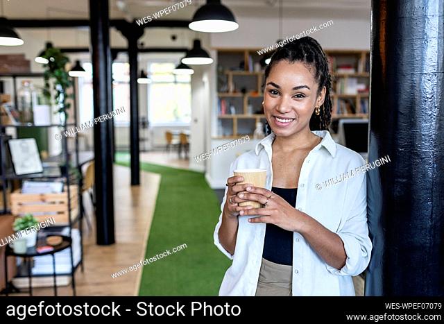 Smiling young businesswoman with disposable coffee cup leaning on column in office