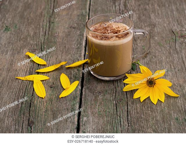 transparent mug with a cappuccino, a yellow flower and petals, a still life, on a wooden table, a subject flowers and drinks