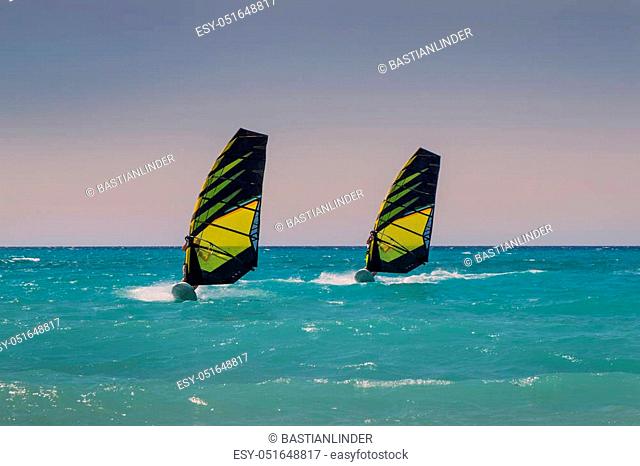 Two windsurfers ride parallel in sea, with similar sails