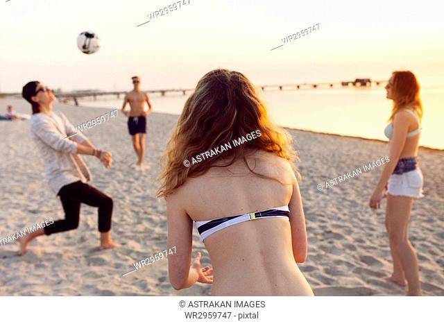Young men, young woman and teenage girl (16-17) playing volleyball on sand at sunset