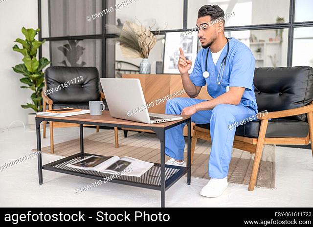 Serious male physician in uniform seated at the laptop talking to his patient via Skype