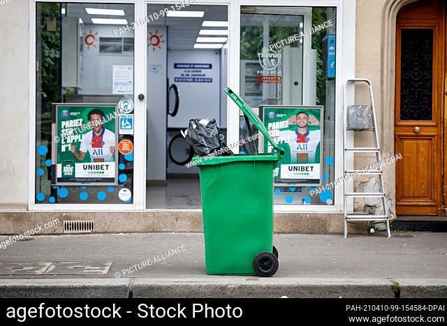 09 April 2021, France, Paris: A garbage can stands in front of a laundromat in the 15th district of Paris. Bulky waste on the sidewalk, overflowing garbage cans