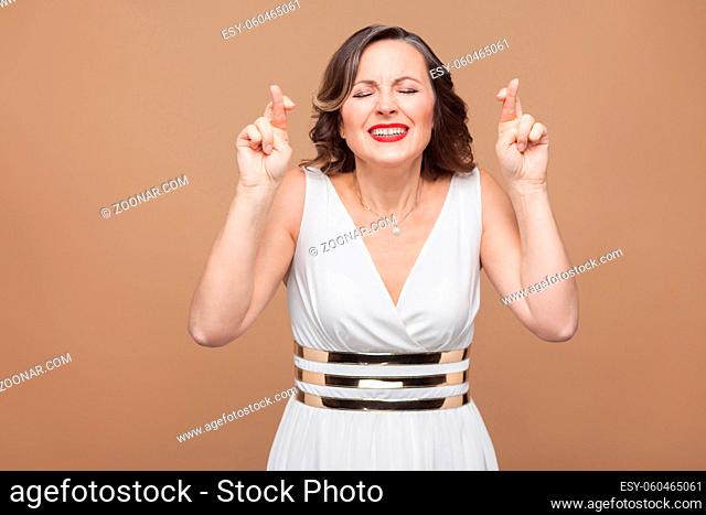 hopeful middle aged woman. Crossed hands and closed eyes. Emotional expressing woman in white dress, red lips and dark curly hairstyle