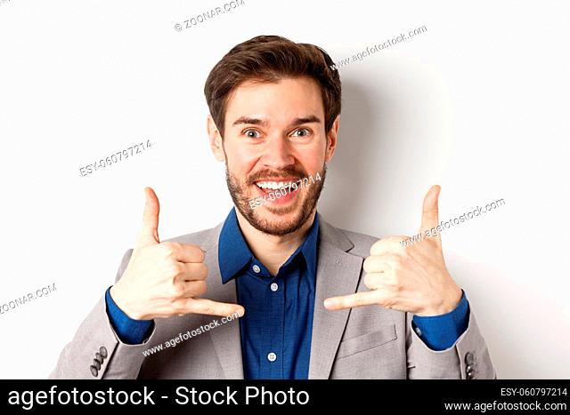 Excited salesman smiling, showing phone signs and asking to give him call, giving his number, standing on white background in suit