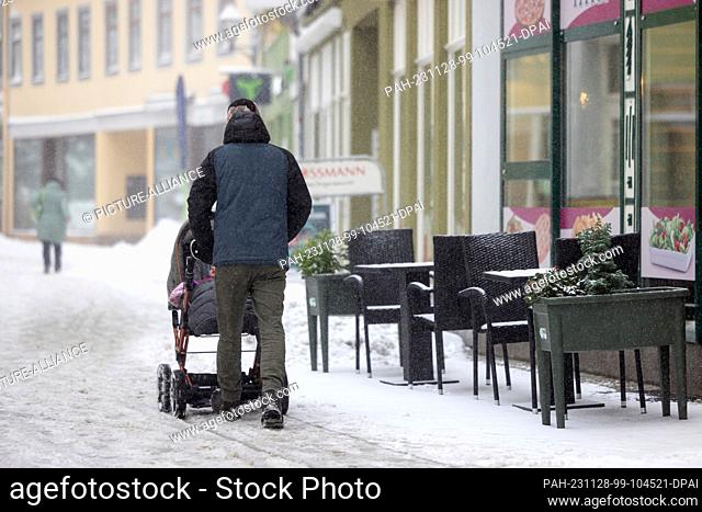 28 November 2023, Thuringia, Ilmenau: A man with a baby carriage walks past snow-covered tables in a restaurant in the city center