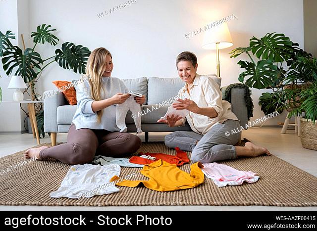 Happy mother and pregnant daughter looking at baby clothes sitting at home