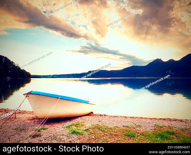Abandoned white paddle boat on Alps lake bank. Evening lake glowing by sunlight. Dramatic and picturesque scene. Mountains in water mirror