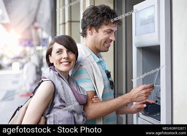 Smiling man withdrawing money from ATM standing by girlfriend