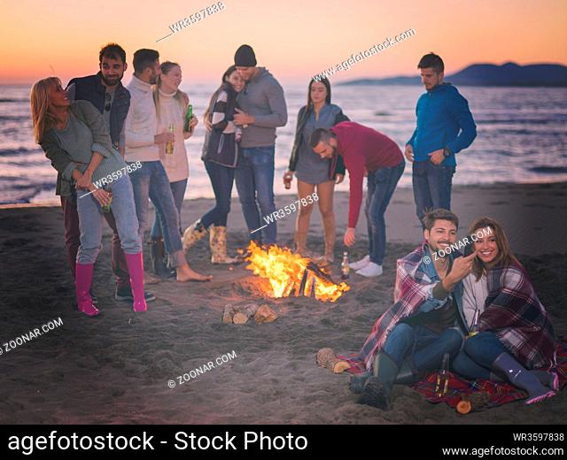 Couple using cell phone during beach party with friends drinking beer and having fun colored filter