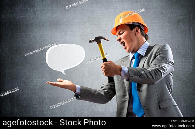Furious businessman going to crash glass speech bubble with hammer. Young handsome man in business suit and safety helmet standing on wall background