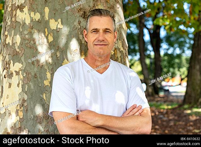 A handsome man leaning on a tree with his arms folded and looking at the camera