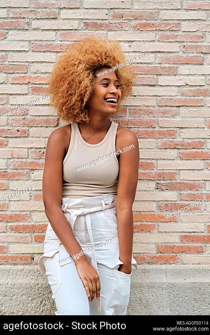 Happy woman with Afro hairstyle looking away while leaning on brick wall